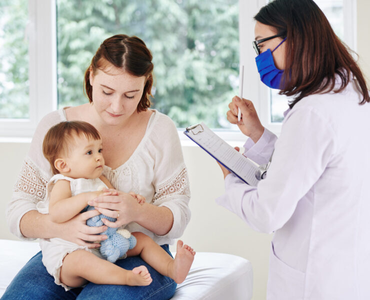 Pediatrician in medical mask talking to baby girl and taking notes in medical card